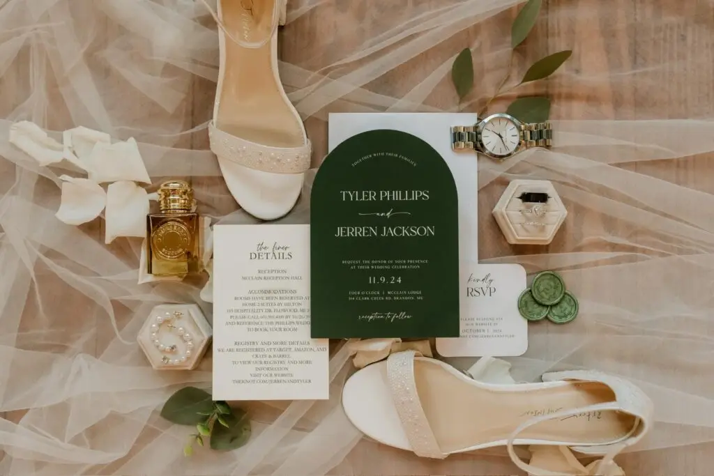 Flatlay of wedding invitations with green accents, bride’s shoes, jewelry, rings, and perfume styled for a Mississippi wedding.