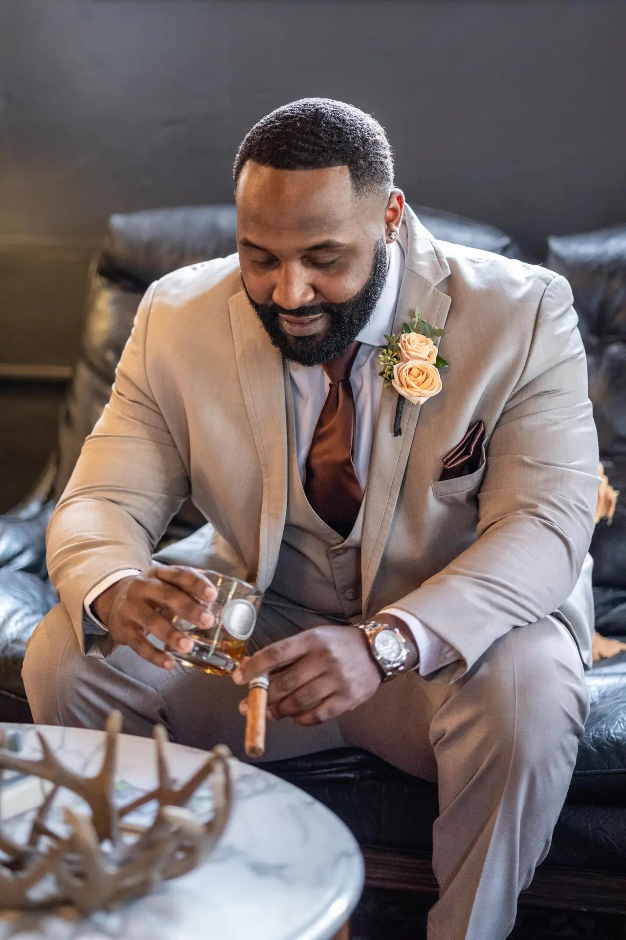 Groom in a beige suit with boutonnière sits with a glass of whiskey and cigar before his Mississippi wedding.