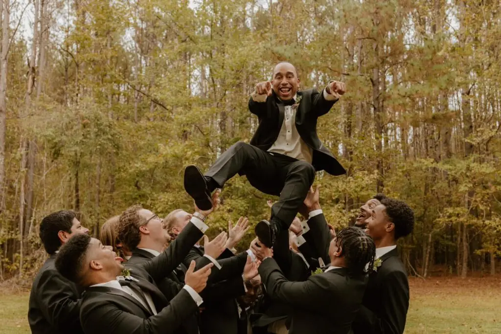 Groomsmen toss groom in the air during outdoor wedding celebration with forest backdrop in Mississippi.